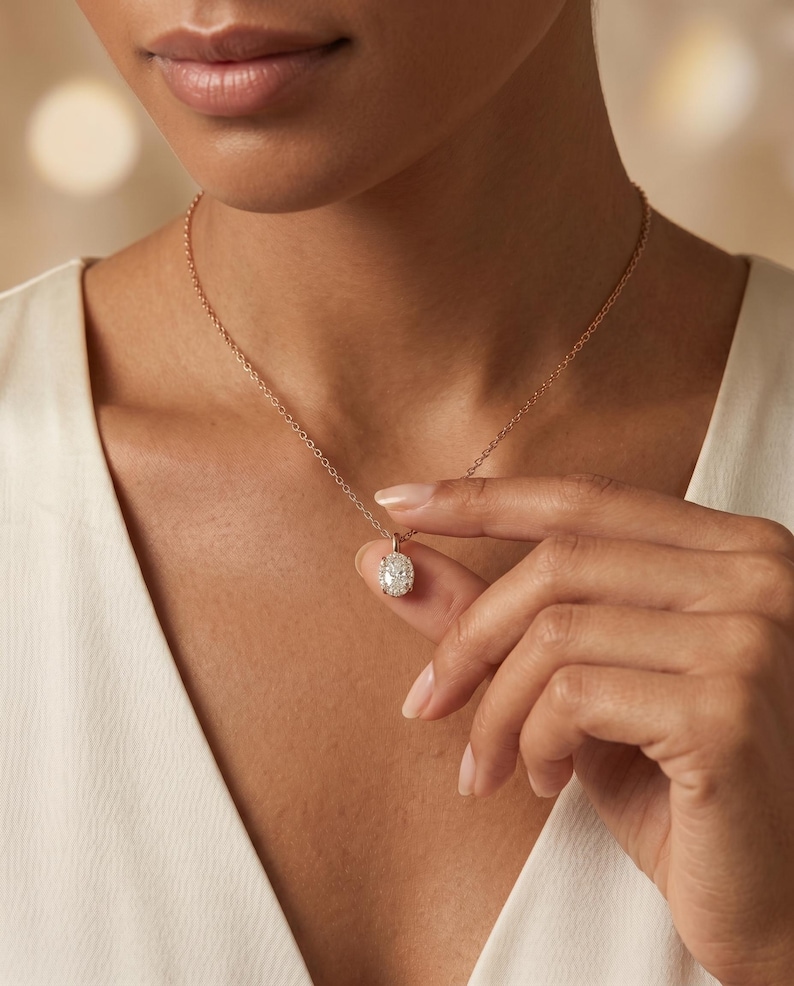 May include: A rose gold necklace with an oval pendant set with small, sparkling stones. The delicate chain is held by a hand. The pendant is a cluster of stones.