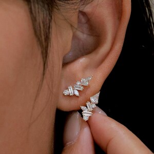 May include: Close-up of two gold earrings with multiple small, clear gemstones. The earrings are worn on an ear, with one earring in the earlobe and the other above it. The gemstones are various shapes, including baguette and round cuts.