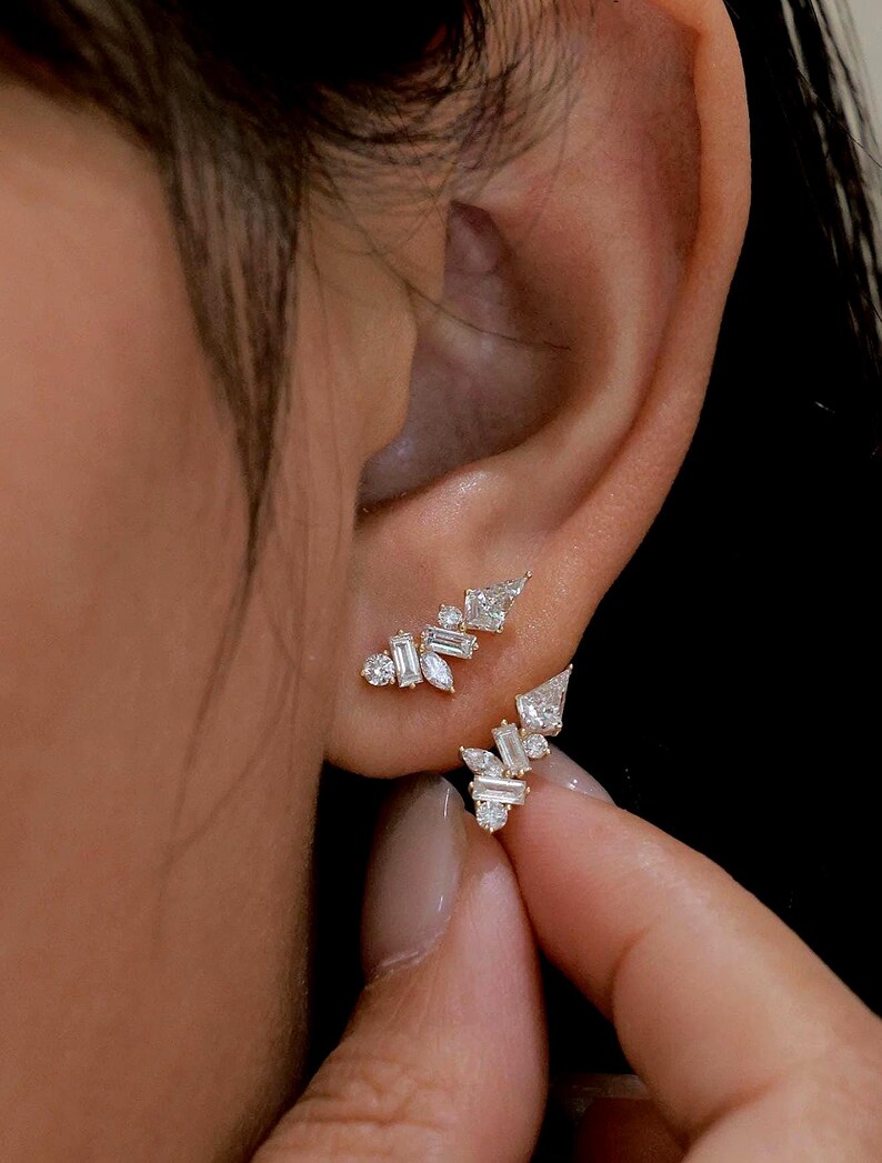 May include: Close-up of two gold earrings with multiple small, clear gemstones. The earrings are worn on an ear, with one earring in the earlobe and the other above it. The gemstones are various shapes, including baguette and round cuts.