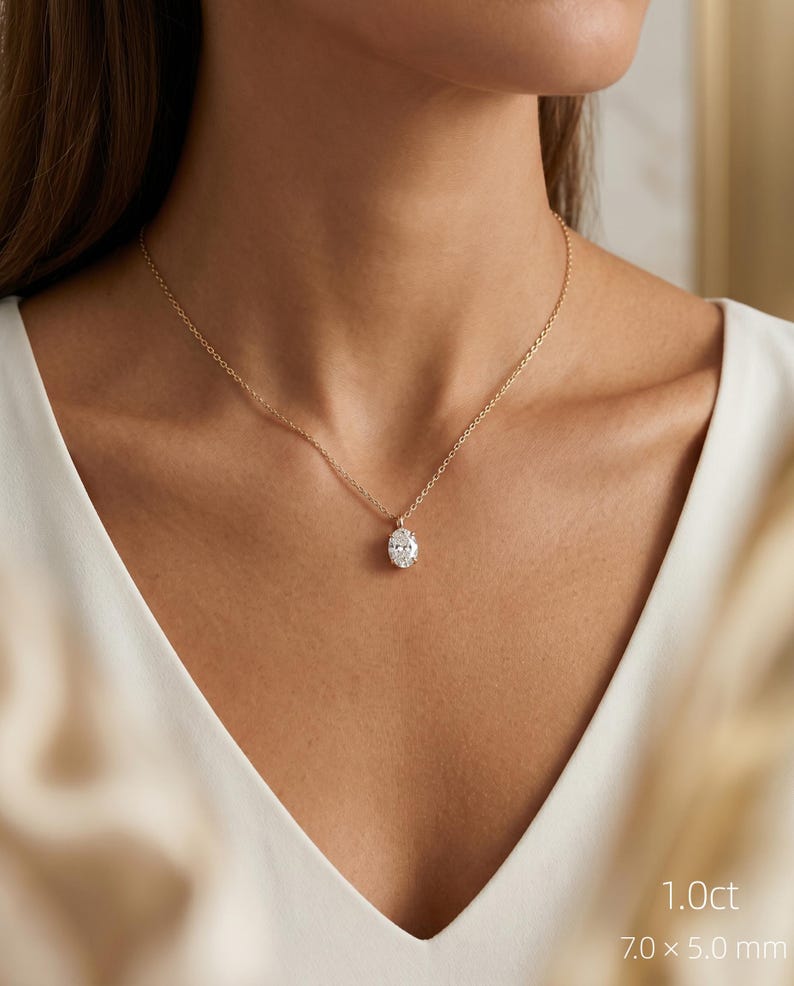 May include: A gold necklace with an oval-cut diamond pendant. The diamond is set in a gold mount and suspended from a fine gold chain. The image text reads "1.0ct" and "7.0 x 5.0 mm". The pendant measures approximately 7.0 mm x 5.0 mm.