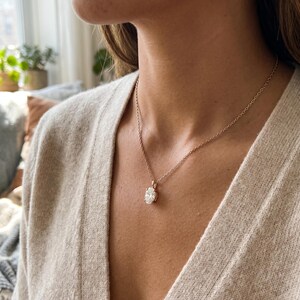 May include: A gold necklace featuring a clear, round gemstone pendant. The pendant is set in a gold bezel and suspended from a fine gold chain. The necklace is worn against a person's skin, with a blurred background.