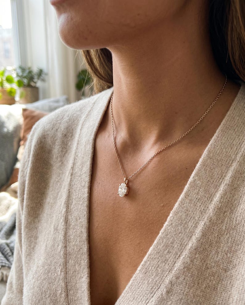May include: A gold necklace featuring a clear, round gemstone pendant. The pendant is set in a gold bezel and suspended from a fine gold chain. The necklace is worn against a person's skin, with a blurred background.