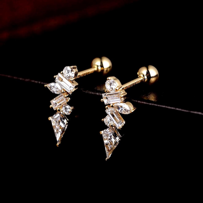 May include: A pair of gold-colored stud earrings with a cluster of clear, sparkling crystals. The crystals are various shapes, including round, baguette, and teardrop, arranged in a delicate, asymmetrical design. The earrings have a post and ball backing.