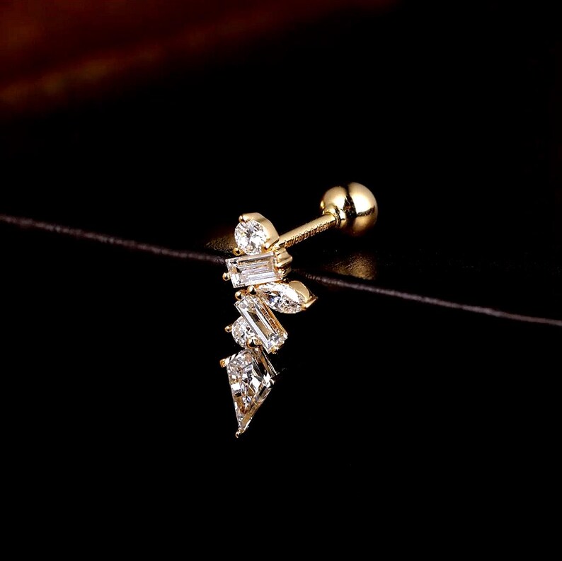 May include: A gold-colored helix earring featuring a cluster of clear, sparkling cubic zirconia stones in various shapes. The design includes round, baguette, and pear-cut stones, creating a delicate, asymmetrical arrangement. The earring has a small, round ball on the end.