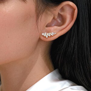 May include: A gold-toned ear climber earring featuring a cluster of clear, sparkling cubic zirconia stones. The design includes various shapes, such as round and baguette-cut stones, arranged in a delicate, asymmetrical pattern. The earring is worn on the ear.