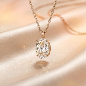 May include: A gold necklace featuring an oval-cut diamond pendant. The diamond is set in a gold bezel and suspended from a fine gold chain. The backdrop is a soft, blurred fabric in a warm, neutral hue, creating a luxurious aesthetic.