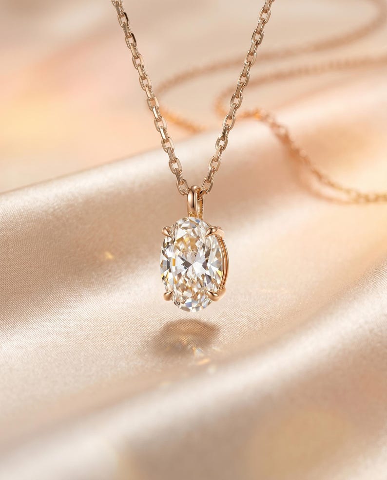 May include: A gold necklace featuring an oval-cut diamond pendant. The diamond is set in a gold bezel and suspended from a fine gold chain. The backdrop is a soft, blurred fabric in a warm, neutral hue, creating a luxurious aesthetic.