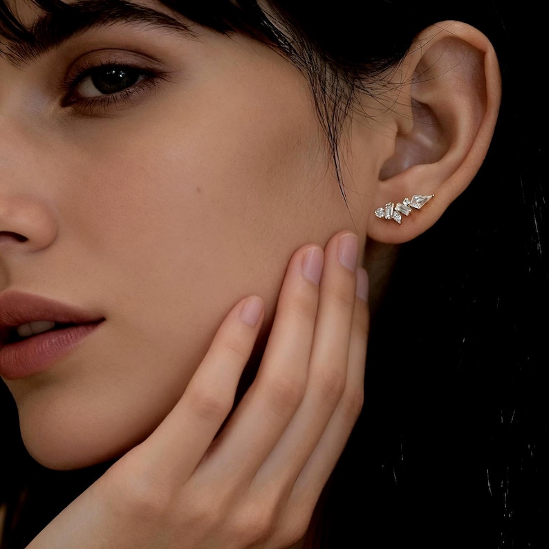 May include: A close-up of a person's ear adorned with a delicate, gold-toned earring. The earring features a cluster of small, sparkling, clear gemstones arranged in an asymmetrical design. The person's hand gently touches their face.