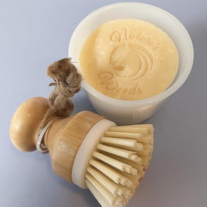 Puede incluir: Una pastilla de jabón redonda de color amarillo claro con las palabras "Nature's Woods" y un diseño de hoja está en un cuenco blanco. Junto a ella, un cepillo de cocina de madera con cerdas naturales y un lazo de cuerda. El fondo es azul claro.