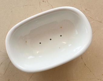 May include: A white, oval soap dish with three drainage holes. The dish has a glossy finish and is set against a neutral background. The design is simple and functional, suitable for a bathroom or kitchen.