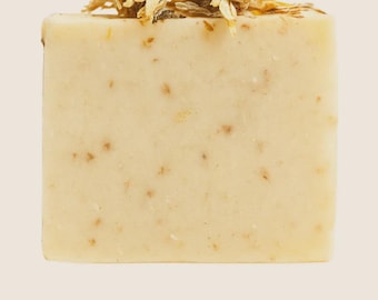 May include: A rectangular bar of handmade soap, light beige with brown flecks. Dried flowers and buds adorn the top. The soap is unscented and natural, ideal for sensitive skin. The bar's dimensions are approximately 8 cm x 5 cm x 3 cm.