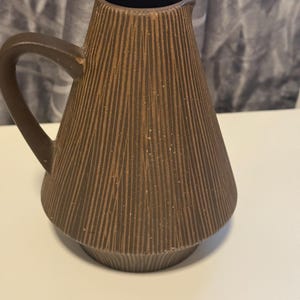 May include: A brown ceramic pitcher with a handle and a textured, vertical-lined design. The pitcher has a wide base that tapers upwards to a narrow opening. The interior is dark brown. The pitcher is likely used for serving beverages.