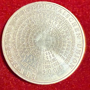 Può includere: Una moneta d'argento con testo a forma circolare. La moneta presenta le parole "DER EUROPÄISCHEN UNION" e "ERWEITERUNG" sul bordo. Il centro presenta date come 1950, 1952, 1958, 1973-1981, 1986-1995 e 2004.