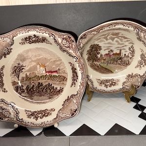 Könnte beinhalten: Zwei antike, cremefarbene Keramikschalen mit braunem Transferdruck. Jede Schale hat einen gewellten Rand und eine zentrale Landschaftsszene mit einem Schloss. Die Ränder sind mit Blumen- und Scrollmustern verziert. Eine Schale steht auf einem goldenen Ständer.