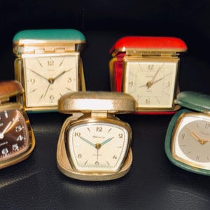 Puede incluir: Una colección de relojes despertadores de viaje vintage en varios colores. Los relojes tienen cajas doradas y esferas de diferentes colores, incluyendo marrón, crema y verde. Los relojes se exhiben sobre una superficie oscura.
