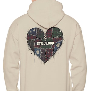 Könnte beinhalten: Beigefarbener Kapuzenpullover mit einem großen, zerbrochenen Herzdesign auf dem Rücken. Das Herz besteht aus verschiedenen Stoffflicken in Blau-, Grün- und Rottönen, mit dem weißen Schriftzug "STILL LOUD". Der Hoodie hat eine Kapuze und lange Ärmel.