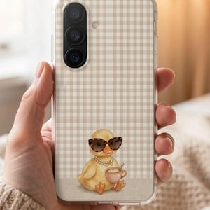 Könnte beinhalten: Transparente Handyhülle mit einem beige-weißen Vichy-Muster. Die Hülle zeigt eine Cartoon-Ente mit Sonnenbrille und Perlenkette, die eine rosa Tasse hält. Die Handyhülle wird in einer Hand gehalten.
