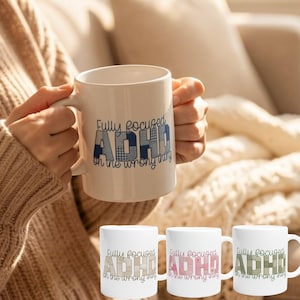 Könnte beinhalten: Eine weiße Keramik-Tasse mit dem Text "Fully focused ADHD on the wrong thing" in blau karierten Buchstaben. Die Tasse wird von einer Person gehalten, die einen beigen Pullover trägt. Drei weitere Tassen mit dem gleichen Design sind sichtbar.