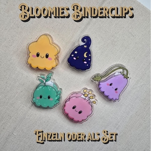Könnte beinhalten: Fünf Acryl-Binderclips mit Comic-Figuren: eine gelbe Wolke, eine dunkelblaue Nachtform mit Mond und Sternen, ein grüner Keimling, eine rosa Blume und eine violette Glockenblume. Text: BLOOMIES BINDERCLIPS und EINZELN ODER ALS SET.