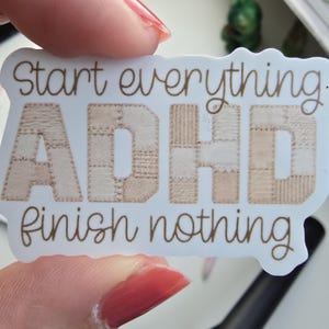 Könnte beinhalten: Ein weißer, konturgestanzter Aufkleber mit dem braunen Text Start everything oben und finish nothing unten. Die zentralen Buchstaben ADHD sind mit einem beigen und hellbraunen Patchwork-Muster gefüllt, das an verschiedene Stoffstrukturen und Nähte erinnert.
