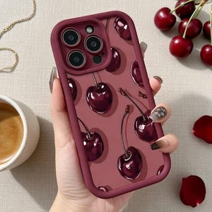 Peut inclure: Coque de téléphone bordeaux ornée d'un motif de cerises rouge foncé avec des tiges vertes. L'étui a un bord surélevé autour des objectifs de l'appareil photo et un bord bordeaux assorti. Le fond est rose.