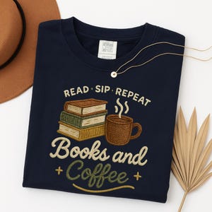 以下が含まれることがあります： 本、コーヒーカップ、そして「READ. SIP. REPEAT.」と「Books and Coffee」の文字が刺繍されたデザインのネイビーブルーのTシャツ。茶色の帽子と金のネックレスが見えます。