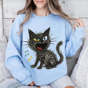 May include: Light blue sweatshirt featuring a whimsical black cat illustration with yellow and blue eyes, an open mouth, and musical notes. The cat is drawn in a scribbled style, giving it a playful appearance. A casual, comfortable top.