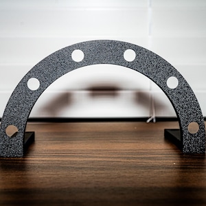 May include: A black, semi-circular metal object with a textured surface and six white circular accents. The object has two black rectangular feet and is displayed on a wooden surface.