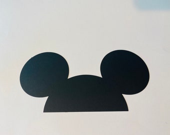 May include: Black paper cutout of Mickey Mouse ears. The ears are rounded and the top of the head is a rounded rectangle.