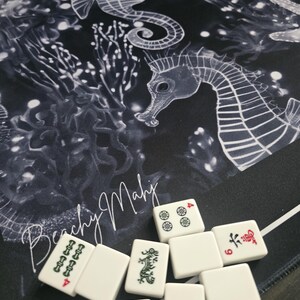 May include: A black and white Mahjong mat with a seahorse design and the text "Beachy Mahj". White Mahjong tiles with red and green markings are scattered on the mat. The tiles display various symbols and numbers.