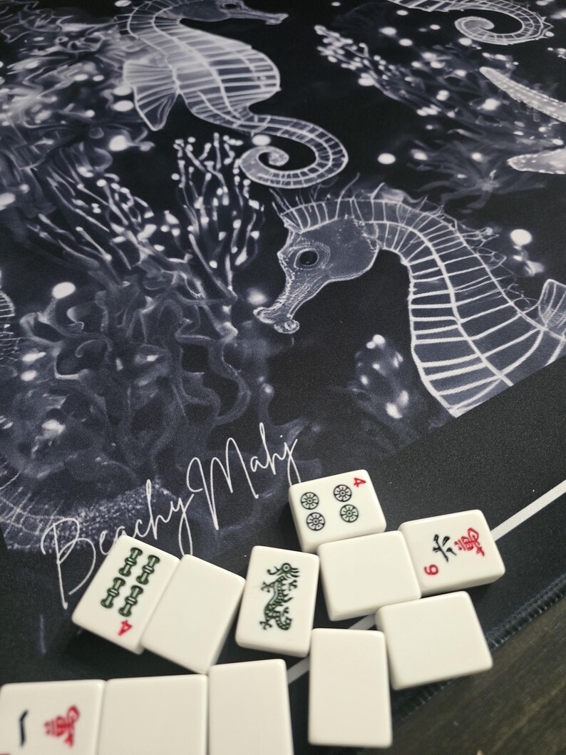 May include: A black and white Mahjong mat with a seahorse design and the text "Beachy Mahj". White Mahjong tiles with red and green markings are scattered on the mat. The tiles display various symbols and numbers.