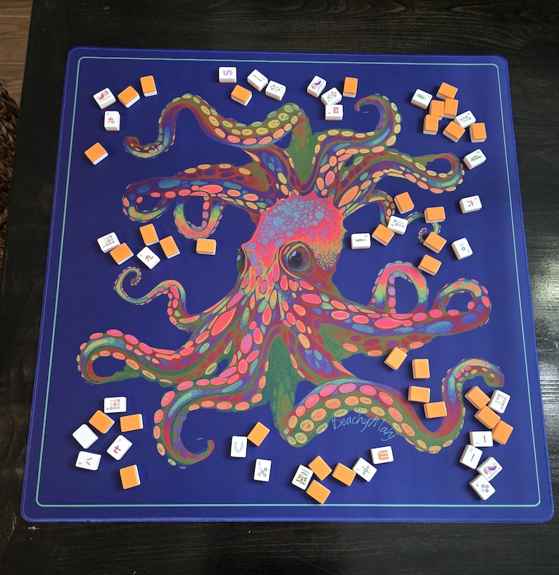 May include: A blue game mat featuring a colorful octopus design with a variety of tentacles. Scattered around the mat are small, rectangular game tiles with white and orange faces. The mat has a teal border and the name "Deachy Mary" is visible.