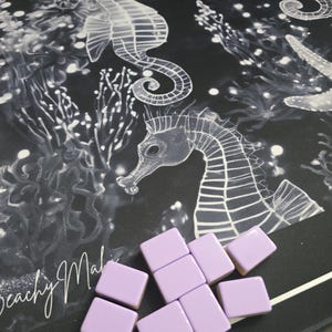 May include: A black and white seahorse and coral design on a mat with light purple square tiles. The mat has the words "Beachy Malibu" in cursive. The tiles are scattered on the mat.