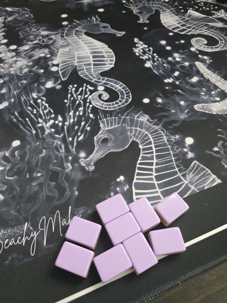 May include: A black and white seahorse and coral design on a mat with light purple square tiles. The mat has the words "Beachy Malibu" in cursive. The tiles are scattered on the mat.