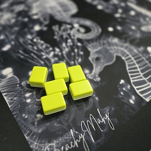 May include: Six small, rectangular, lime green tiles with rounded edges are arranged on a black surface. The surface features a seahorse illustration and the handwritten text "Beachy Mahj". The tiles are the focal point against the detailed background.