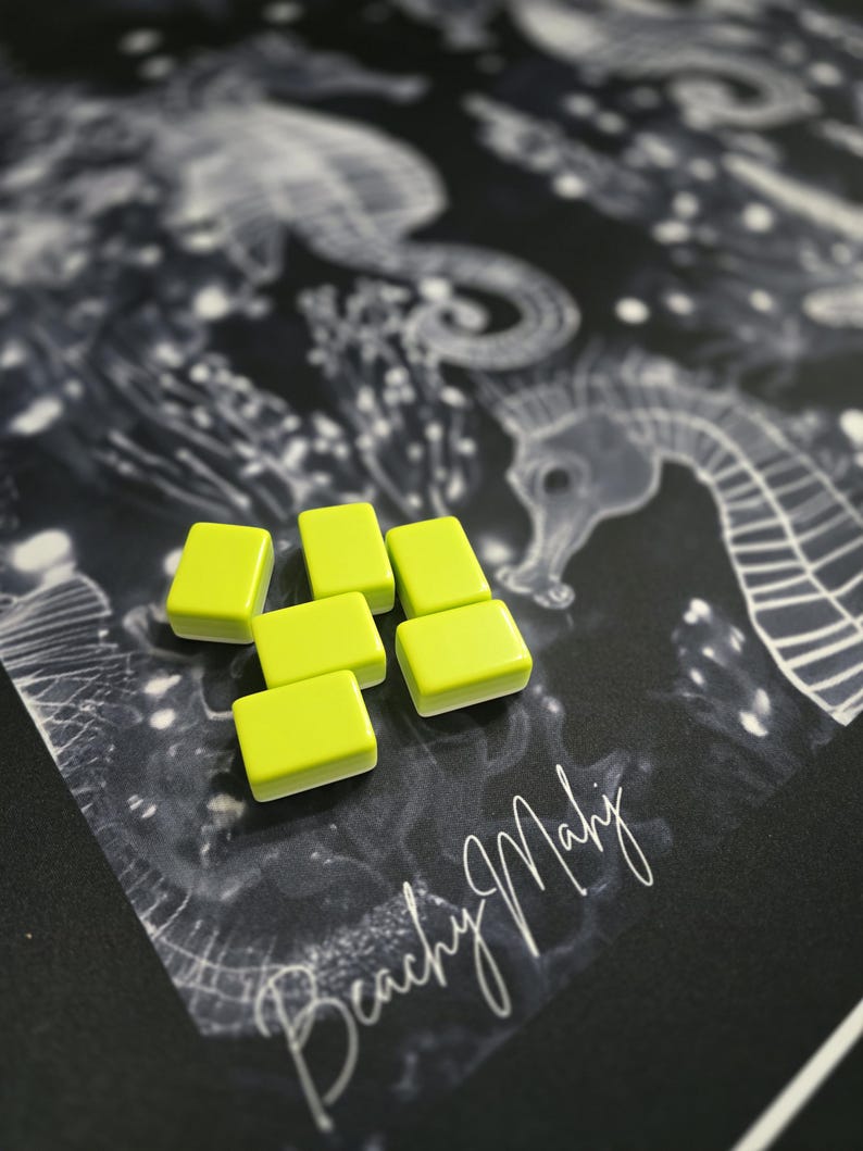 May include: Six small, rectangular, lime green tiles with rounded edges are arranged on a black surface. The surface features a seahorse illustration and the handwritten text "Beachy Mahj". The tiles are the focal point against the detailed background.