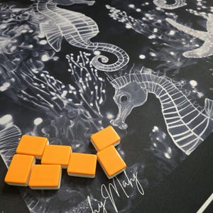 May include: A black game mat with a white seahorse and starfish design. Orange and white game tiles are scattered on the mat. The words "Beachy Mahj" are written in white script on the mat.
