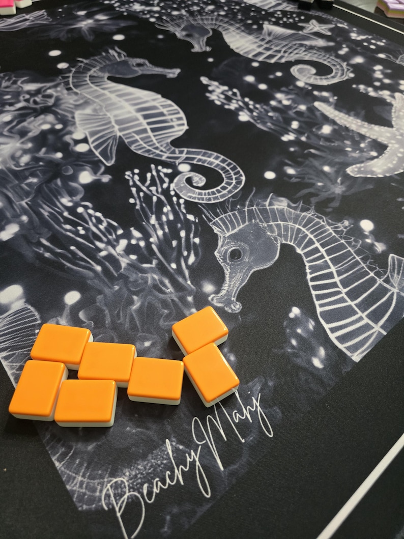 May include: A black game mat with a white seahorse and starfish design. Orange and white game tiles are scattered on the mat. The words "Beachy Mahj" are written in white script on the mat.