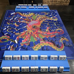 May include: A complete Mahjong set with blue tiles and racks, arranged on a blue mat featuring a colorful octopus design. The tiles are white with blue backs. The set includes racks for holding the tiles during gameplay.