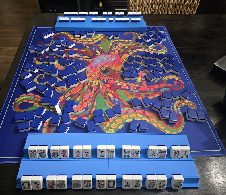 May include: A complete Mahjong set with blue tiles and racks, arranged on a blue mat featuring a colorful octopus design. The tiles are white with blue backs. The set includes racks for holding the tiles during gameplay.