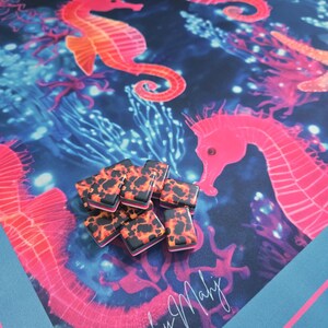 May include: A vibrant beach towel featuring a colorful underwater scene with seahorses in shades of pink and orange against a dark blue background. A pile of black and orange patterned rectangular objects sits on the towel. The towel has the text "Beachy Mahy".