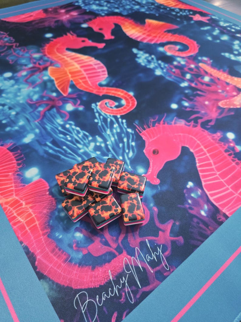 May include: A vibrant beach towel featuring a colorful underwater scene with seahorses in shades of pink and orange against a dark blue background. A pile of black and orange patterned rectangular objects sits on the towel. The towel has the text "Beachy Mahy".