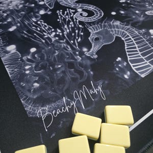 May include: A black and white seahorse-themed game mat with the text "Beachy Mahj" in a cursive font. Several light yellow game tiles are arranged on the mat. The mat features seahorse illustrations and aquatic plant designs.