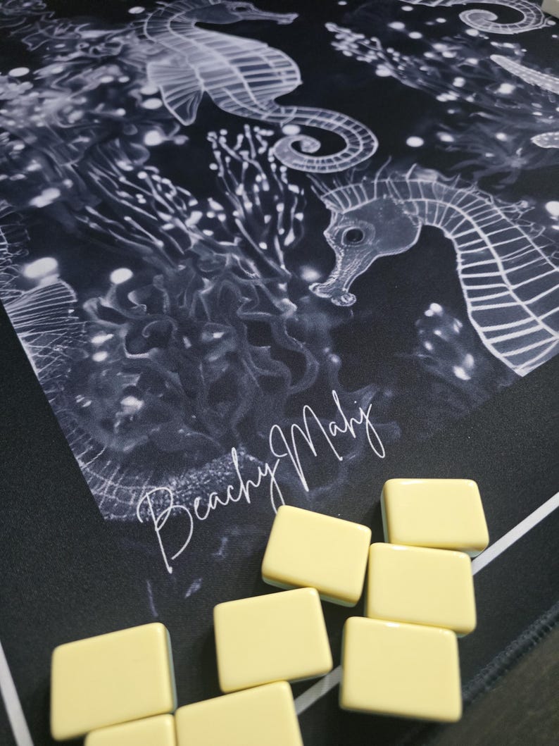 May include: A black and white seahorse-themed game mat with the text "Beachy Mahj" in a cursive font. Several light yellow game tiles are arranged on the mat. The mat features seahorse illustrations and aquatic plant designs.