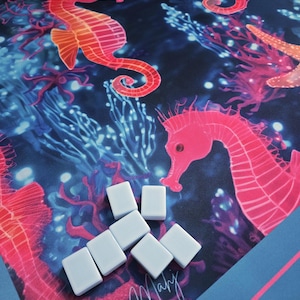 May include: A colorful, ocean-themed game mat featuring seahorses, coral, and starfish in shades of pink, orange, and blue. White game pieces are scattered across the mat. The words "Beachy Mahy" are written in cursive.