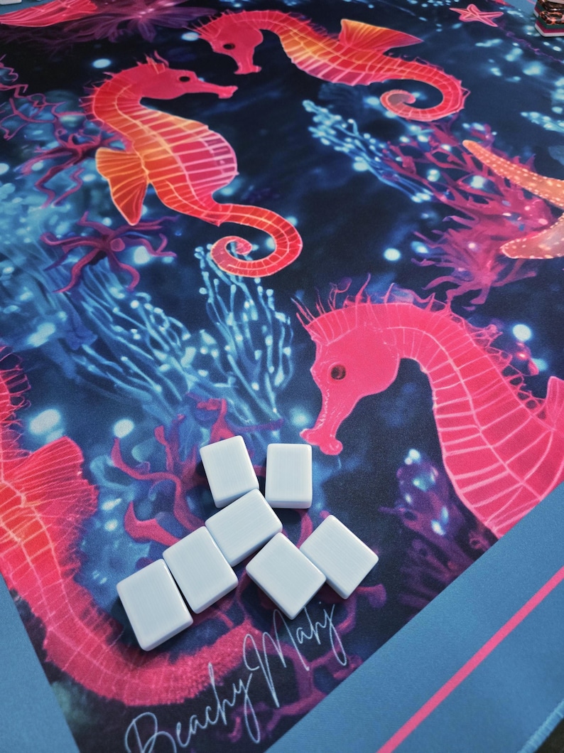May include: A colorful, ocean-themed game mat featuring seahorses, coral, and starfish in shades of pink, orange, and blue. White game pieces are scattered across the mat. The words "Beachy Mahy" are written in cursive.
