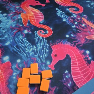 May include: A vibrant, marine-themed game mat featuring coral, seahorses, and other sea life in shades of pink, orange, and blue. Includes a set of orange game tiles. The mat has a blue border and a signature in the corner.