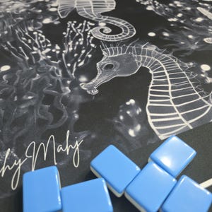 May include: A black and white seahorse and coral design on a fabric surface. Light blue and white Mahjong tiles are scattered across the surface. The text "by Mahj" is visible in white script.