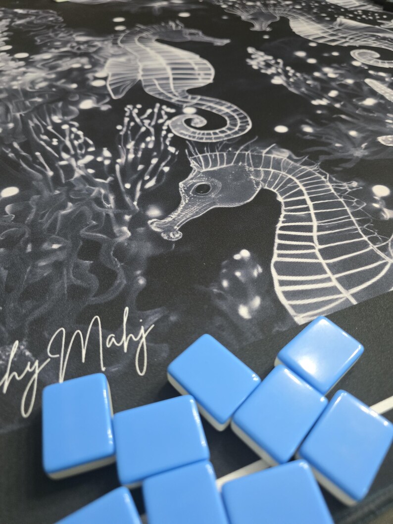 May include: A black and white seahorse and coral design on a fabric surface. Light blue and white Mahjong tiles are scattered across the surface. The text "by Mahj" is visible in white script.