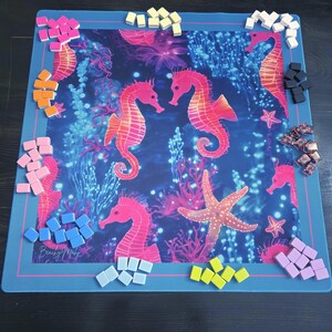 May include: A blue game mat featuring a vibrant underwater scene with seahorses, coral, and a starfish. Various colored game pieces surround the mat. The mat has a pink border and the text "Beach My Way".